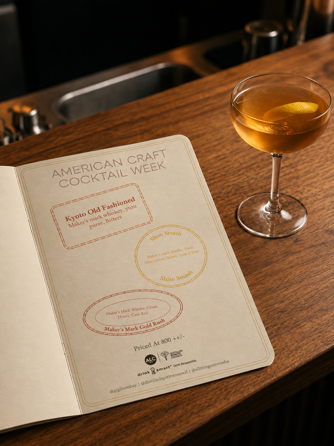 Open American Craft Cocktail Week menu on a wooden bar beside a coupe cocktail with a lemon twist
